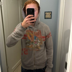 Grå hoodie med tryck från D&G - Grå hoodie från Dolce & Gabbana med färgglatt tryck och röda ränder på ärmarna. Dragkedja framtill, huva och fickor på sidorna.