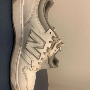 Vita New Balance sneakers med grå detaljer - Snygga vita New Balance sneakers med grå detaljer och klassisk N-logga på sidan. Skorna har snörning, rund tå och platt sula. Ovandelen är i skinn och syntet, vilket ger en clean och sportig look. Perfekta för dig som gillar stilrena och bekväma sneakers.