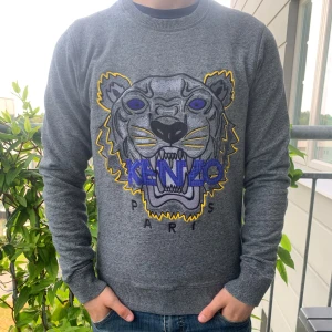 Grå Kenzo sweatshirt med tigertryck - Säljer en grå sweatshirt från Kenzo med ikoniskt broderat tigerhuvud i svart, lila, gult och blått framtill. Tröjan har rund hals, långa ärmar och ribbade muddar. Perfekt statement-plagg för dig som gillar streetstyle och vill sticka ut. Passar dig som är mellan 16-175 ish