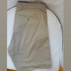 Beige shorts från Gant W33 - Snygga beige shorts från GANT i klassisk chino-stil samt i regular fit. De har bälteshällor, fickor fram och bak, samt en diskret broderad Gant-logga ovanför bakfickan. Stilrena och lätta att matcha med det mesta. Shortsen är i bra skick och har även kvar sina extra knappar (se sista bilden). 