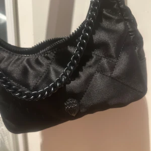Svart quiltad axelväska Kurt Geiger - Snygg svart quiltad axelväska från Kurt Geiger med kedjedetalj och dragkedja upptill. Väskan har en glansig finish och är tillverkad i syntetmaterial. Perfekt storlek för det viktigaste och med cool detalj av Kurt Geigers ikoniska örn-logga framtill.