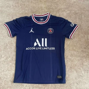 PSG matchtröja marinblå Jordan - Marinblå PSG fotbollströja från Jordan med klubbmärke på bröstet, vit sponsorlogga och röda, vita och blå detaljer på krage och ärmslut. Kortärmad modell i lätt och ventilerande material, perfekt för fotbollsplanen eller att bära till vardags.