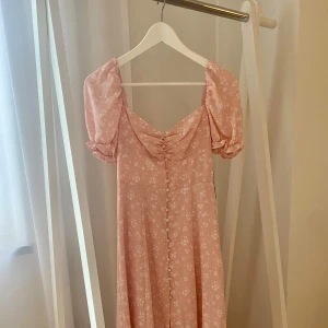 Rosa blommig långklänning från NA-KD storlek 34 - Supersöt långklänning i ljusrosa från NA-KD med vita småblommor, puffärmar och knappar framtill. Klänningen är helt ny med lappar kvar. Storlek 34.