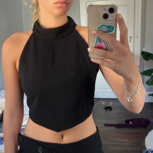 Svart croppad halterneck topp Zara - Snygg svart croppad halterneck topp från Zara med hög krage och ärmlös design. Toppen har en stilren, spetsig nederkant och stängs med knapp i nacken. 