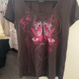 Brun t-shirt med cowboy boots från Esprit - Cool brun t-shirt från Esprit med tryck av rosa och röda cowboy boots framtill. T-shirten har rund halsringning och korta ärmar. Materialet är mjuk bomull, perfekt för en avslappnad look med en touch av western vibes.