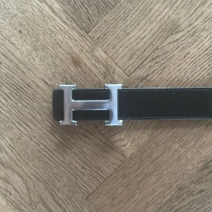 H-bälte vänbart - Säljer ett vändbart H-bälte i skinn med ikoniskt H-spänne i silver. Ena sidan är svart och den andra är blå, båda med kontrastsömmar. Perfekt accessoar för att lyfta din outfit med en lyxig touch.