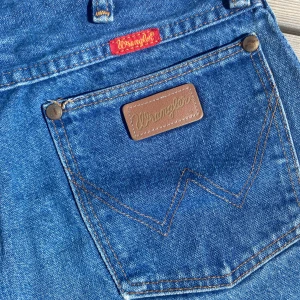 Wrangler högmidjade jeansshorts - Snygga blå jeansshorts från Wrangler med hög midja och fransiga benslut. Klassisk femficksmodell med bruna detaljer och logga på bakfickan. Perfekta för sommaren och ger en avslappnad vintagekänsla. Endast använda ett fåtal gånger och inga defekter förekommer! 💖