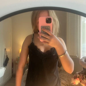 Svart linnetopp med spetsdetaljer - Säljer en svart linnetopp med tunna axelband och snygga spetsdetaljer längs urringningen. Toppen har en mjuk och följsam känsla, perfekt att styla med jeans eller kjol. Enkel men ändå elegant look.