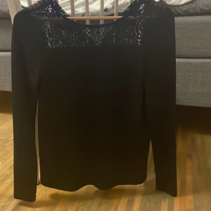 Svart långärmad topp med spetsdetalj - Snygg svart långärmad topp med vacker spets upptill över axlar och bröst. Spetsen har ett detaljerat mönster som ger en elegant känsla. Perfekt för dig som vill ha en stilren men ändå unik look. Materialet känns mjukt och följsamt.