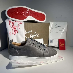 Grå mockasneakers från Christian Louboutin - Snygga grå sneakers i mocka från Christian Louboutin med klassisk röd sula och vita kanter. Skorna har rund tå, platt sula och grå snörning. Insidan är fodrad i beige skinn för extra komfort. Perfekt för dig som vill sticka ut med exklusiv stil.