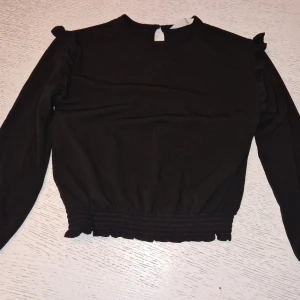 Svart långärmad blus med volang H&M - Svart långärmad blus från H&M med volangdetaljer på axlarna och smockad resår i midjan. Enkel och stilren modell som passar perfekt till jeans eller kjol. Rund halsringning och knapp i nacken för enkel påklädning.