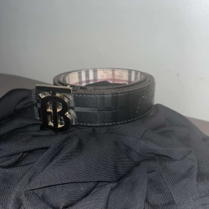 Burberry skärp box, dustbag mm ingår - Snyggt Burberry-bälte i svart och beige med klassiskt rutigt mönster och ikoniskt B-spänne i metall. Bältet är gjort i skinn och har både mörka och ljusa toner. Perfekt accessoar för att lyfta din outfit med en lyxig touch.