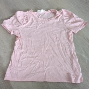 Ljusrosa t-shirt från Kappahl - Söt ljusrosa t-shirt från Kappahl i 100% bomull. Modellen har korta puffärmar och en klassisk rund halsringning. Perfekt för dig som gillar en enkel men gullig stil.