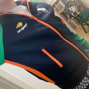 Ralph Lauren US Open jacka blå/grön - Snygg track jacket från Ralph Lauren med US Open-tryck. Jackan är marinblå med gröna ärmar och orange detaljer längs dragkedja och ärmar. Stort vitt polo logga på ryggen och mindre logga fram. Perfekt för dig som gillar sportig och färgstark stil. Helt oänvänd.