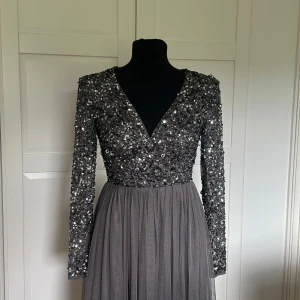 Grålila glittrig midiklänning från Maya Deluxe - Säljer en superfin grålila, midilång klänning från Maya Deluxe med glittrig paljettöverdel och v-ringning. Klänningen har långärmade ärmar och en plisserad tyllkjol. Tyvärr saknas några paljetter på baksidan av vänster ärm, men det går säkert att åtgärda med liknande paljetter för den som är händig! Klänningen är annars helt ny och har aldrig använts. Storlek XS 