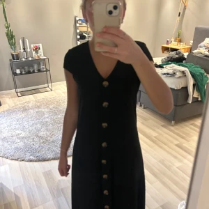 Svart ribbad klänning med knappar - Svart ribbad klänning med v-ringning och stora dekorativa knappar framtill. Klänningen har korta ärmar och en något utställd passform som slutar under knäet. Perfekt för dig som gillar enkel men stilren design.