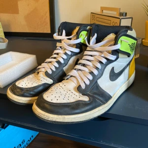 Nike Air Jordans med svart, vit och gul detalj - Säljer ett par Nike Air Jordans med svart och vit ovandel, svarta detaljer och gula inslag vid hälen. Skorna har snörning, rund tå och platt sula. Klassisk Nike-logga på sidan och vinge på ankeln. Perfekta för dig som gillar streetwear.