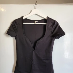 Svart topp med djup v-ringning - En svart, tight topp med korta ärmar och djup v-ringning. Tillverkad i ett stretchigt, mjukt material som sitter figurnära. Storlek S. Endast provad. 