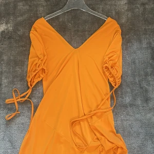 Orange långklänning med puffärmar från Zara - Säljer en orange långklänning från Zara med v-ringning och puffiga kortärmar med knytband. Klänningen har en lös passform och är tillverkad i ett mjukt, lätt material. Perfekt för sommarens alla tillfällen!