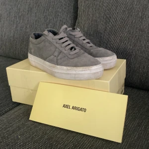 Grå sneakers från Axel Arigato storlek 42 - Säljer ett par stilrena grå sneakers från Axel Arigato i mocka med rund tå och platt sula. Skorna har klassisk snörning och en vit sula som ger en clean look. Perfekta för dig som gillar minimalistisk stil.
