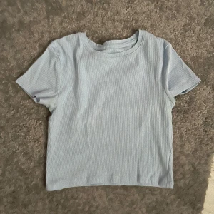 Ljusblå ribbad t-shirt - Säljer en ljusblå ribbad t-shirt med korta ärmar och rund halsringning. Perfekt för en enkel och stilren look. Croppad tröja så ganska kort
