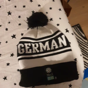 Svartvit mössa med texten GERMANY och FIFA Women's World Cup-märke - Snygg svartvit mössa med stor text 'GERMANY' och svart tofs på toppen. Mössan har ett officiellt FIFA Women's World Cup 2023-märke framtill och bred svart uppvikt kant. Perfekt för fotbollsfans!