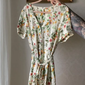 Blommig omlottklänning med knytband - Söt kortärmad klänning med blommigt mönster i grönt, rosa och gult på vit botten. Klänningen har v-ringning, knytband i midjan och en lätt, luftig känsla – perfekt för varma dagar.