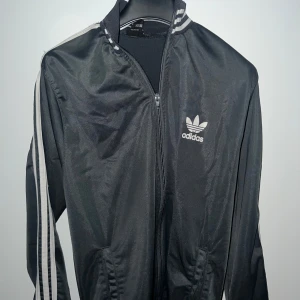 Svart Adidas track jacket med vita ränder - Klassisk svart Adidas track jacket med vita ränder längs ärmarna och vit Adidas-logga på bröstet. Jackan har dragkedja framtill och ribbade muddar vid krage och ärmslut. Perfekt för en sportig och avslappnad stil.
