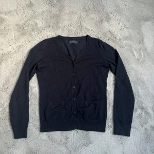 Svart kofta från Stockh LM - En klassisk kofta från Stockh LM med v-ringning och knäppning framtill. Tröjan har långa ärmar, två små fickor framtill och är tillverkad i ett tunt, mjukt material. Perfekt att slänga över en topp för en enkel look.