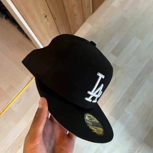 Svart LA-keps från New Era 59FIFTY - Snygg svart keps från New Era 59FIFTY med LA-logga broderad framtill i vitt. Klassisk rak skärm och broderad MLB-logga baktill. Perfekt för dig som gillar streetwear och sportig stil.
