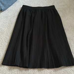 Svart plisserad kjol från Lindex - Svart plisserad kjol från Lindex med klassisk midja och vackert fall. Kjolen är lång och har en elegant, tidlös look som passar perfekt till många olika stilar. Tillverkad i ett lätt och följsamt material.