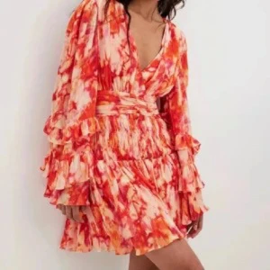 Sanna Jörnvik Nakd klänning - Säljer en superfin kortklänning från Sanna Jörnviks kollektion med NA-KD i storlek 32. Klänningen har ett färgstarkt mönster i rött, orange och rosa, v-ringning, volangdetaljer och långa ärmar. Perfekt för dig som vill ha den perfekta sommarklänningen.