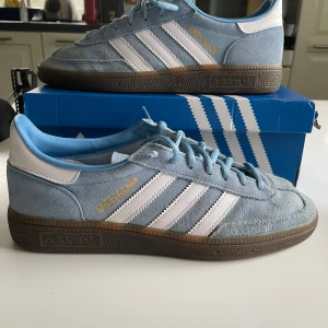 Adidas Handball Spezial ljusblå sneakers - Säljer ett par Adidas Handball Spezial sneakers i ljusblå mocka med vita detaljer och klassiska tre ränder på sidan. Skorna har snörning, brun platt sula och guldtext på sidan. Perfekta för dig som gillar retrostil och sportiga vibes.