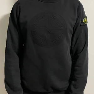 Svart sweatshirt från Stone Island - Säljer en svart sweatshirt från Stone Island med stort broderat logomärke på bröstet och klassisk patch på ärmen. Tröjan är i bomull och har rund hals samt långa ärmar. Tröjan är i storlek XL men har passform för M! 