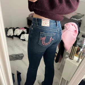 True Religion Jeansbyxor Becca Mid Rise Bootcut med rosa broderi - Säljer ett par blå jeansbyxor från True Religion med bootcut-modell och midwaist. Bakfickan har ett snyggt rosa broderat mönster. Klassisk femficksdesign och bekväm passform, perfekta till vardags.
