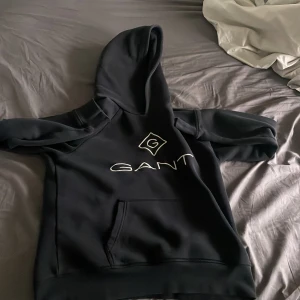 Svart hoodie från GANT - Säljer en svart hoodie från GANT med vit logga på bröstet. Tröjan har huva och en stor magficka framtill. Perfekt för en avslappnad och stilren look.