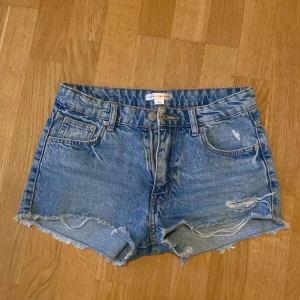Blå jeansshorts från Gina Tricot - Säljer ett par klassiska blå jeansshorts från Gina Tricot med råa kanter och slitningar framtill. Shortsen har fem fickor och knappgylf. Perfekta för sommaren och ger en avslappnad look.