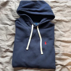 Mörkblå Ralph Lauren hoodie - Säljer en mörkblå hoodie från Polo Ralph Lauren med vit dragsko och klassisk röd logga broderad på bröstet. Tröjan har känguruficka och mjukt material, perfekt för en avslappnad stil.