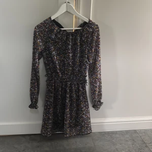 Blommig kortklänning med lång ärm - Söt kortklänning med småblommigt mönster i flera färger på svart botten. Klänningen har långa ärmar, markerad midja med resår och lätt volang vid ärmslut. Perfekt för dig som gillar romantisk stil.