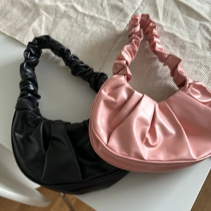 Rynkad axelväska i rosa och svart - Två trendiga axelväskor med rynkat axelband, en i ljusrosa och en i svart. Väskorna har en mjuk, rundad form och är tillverkade i ett slätt syntetmaterial som ger en modern look. Perfekt accessoar för att lyfta din outfit. Båda för 120 eller bud 