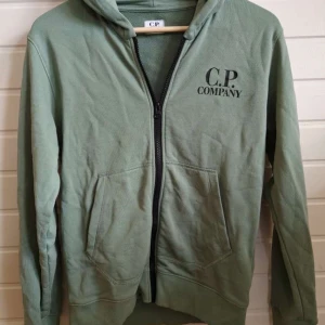 Grön hoodie med glasögon från C.P. Company - Snygg grön hoodie från C.P. Company med dragkedja och huva med inbyggda glasögon. Jackan har två fickor framtill och logga på bröstet. Perfekt för dig som gillar streetwear och unika detaljer. Knappen som man spänner fast glasögon med har lossnad men det är bara att sy fast. Priset går alltid att diskutera!