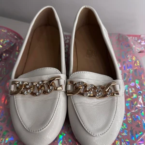 Vita loafers med guldkedja och strass - Vita loafers med dekorativ guldfärgad kedja och glittrande strassdetaljer framtill. Skorna har rund tå och platt sula, i storlek 36. Har använt de två gånger. 
