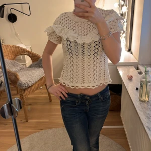 Vit virkad blus med volang - Superfin vit virkad blus med korta ärmar och stor volang runt axlarna. Blusen har ett romantiskt mönster och är croppad med söt kant nertill. Perfekt för dig som gillar en feminin och bohemisk stil.