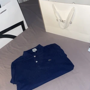 Mörkblå pikétröja från Lacoste - Säljer en klassisk mörkblå pikétröja från Lacoste med den ikoniska krokodilloggan broderad på bröstet. Tröjan har krage, knappar framtill och är tillverkad i bomull. Perfekt för en stilren och sportig look.
