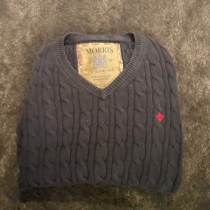 Mörkblå kabelstickad tröja från Morris - Säljer en mörkblå kabelstickad tröja från Morris med v-ringad hals och diskret röd broderad logga på bröstet. Tröjan har långa ärmar och ribbade muddar vid ärmslut och nederkant. Perfekt för finare/exklusiv klädsel