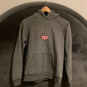 Grå hoodie från GANT - Säljer en grå hoodie från GANT med broderad logga och sköld med stjärnor och ränder på bröstet. Tröjan har huva och en stor magficka framtill. Perfekt för en avslappnad stil. Nypris 599kr
