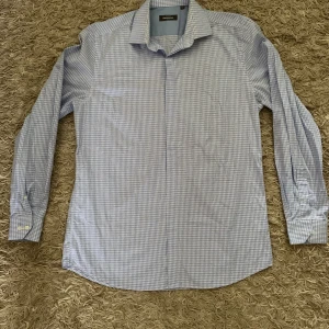 Blårutig skjorta från Dressmann - Snygg blårutig skjorta från Dressmann med klassisk krage och långa ärmar. Skjortan har knappar framtill och är tillverkad i ett lätt och bekvämt material. Perfekt för en stilren look.