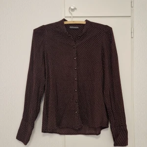 Vinröd prickig blus från Nygårdsanna - Säljer en vinröd blus från Nygårdsanna med små vita prickar. Blusen har långa ärmar, knappar framtill och vid ärmslut samt en lätt v-ringad hals. Perfekt för dig som gillar klassiska mönster och mjuka material.