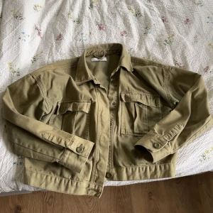 Olivgrön jeansjacka  - Säljer en olivgrön jeansjacka. Jackan har klassisk krage, två bröstfickor med lock och knappar framtill. Modellen är något croppad och har långa ärmar med knapp vid ärmslut. Jag har vanligtvis strl M och jackan passar jättebra på mig. Skriv gärna vid frågor🩷