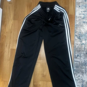 Svarta träningsbyxor från Adidas med vita ränder - Svarta Adidas träningsbyxor med klassiska vita ränder längs sidorna och elastisk midja. Perfekta för sport eller chill. Adidas-logga (helt svart) på vänster ben och raka ben för en avslappnad look.
